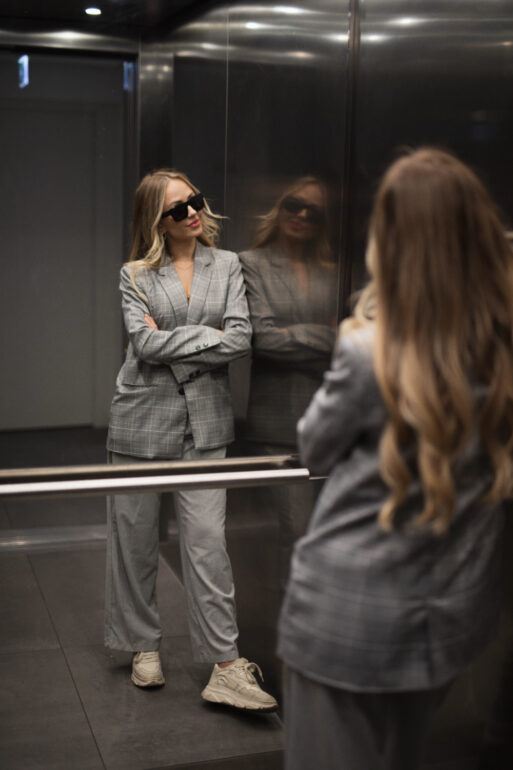 Женщина в сером клетчатом костюме и кроссовках стоит в лифте, отражаясь в зеркале, создавая энергию зеркала в лифте.